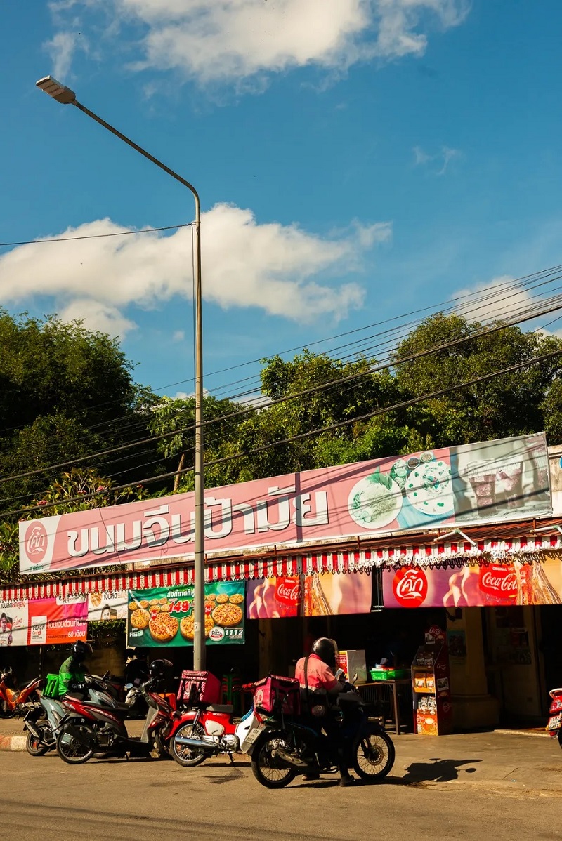 Người đi xe máy đậu bên ngoài chợ quảng cáo đồ ăn Thái dưới bầu trời xanh.