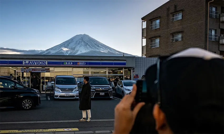 Một khách du lịch tạo dáng trước cửa hàng tiện lợi Lawson với nền là núi Phú Sĩ.